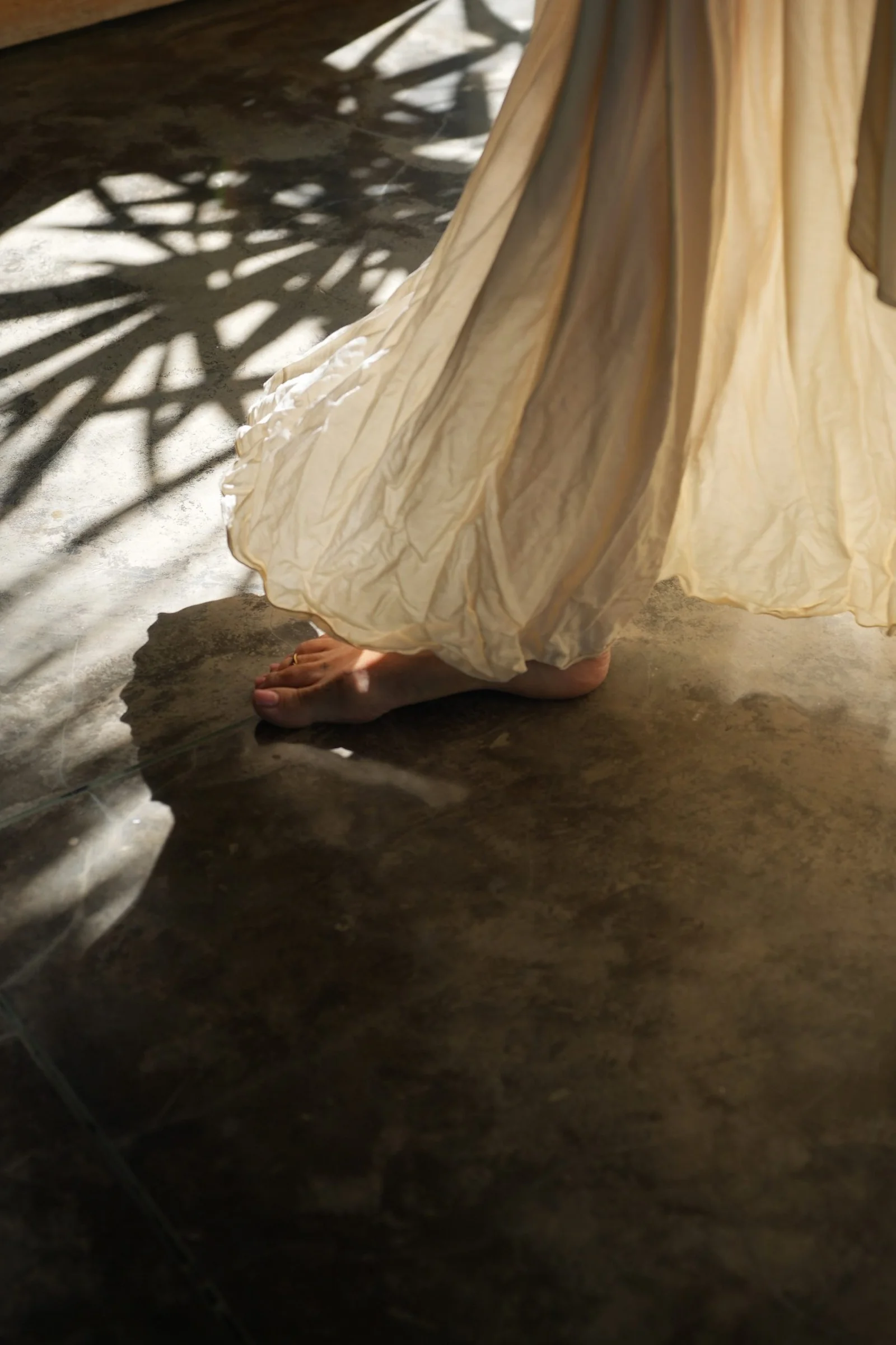 Woman in a flowing cream dress walking through palm shadows on a concrete floor
