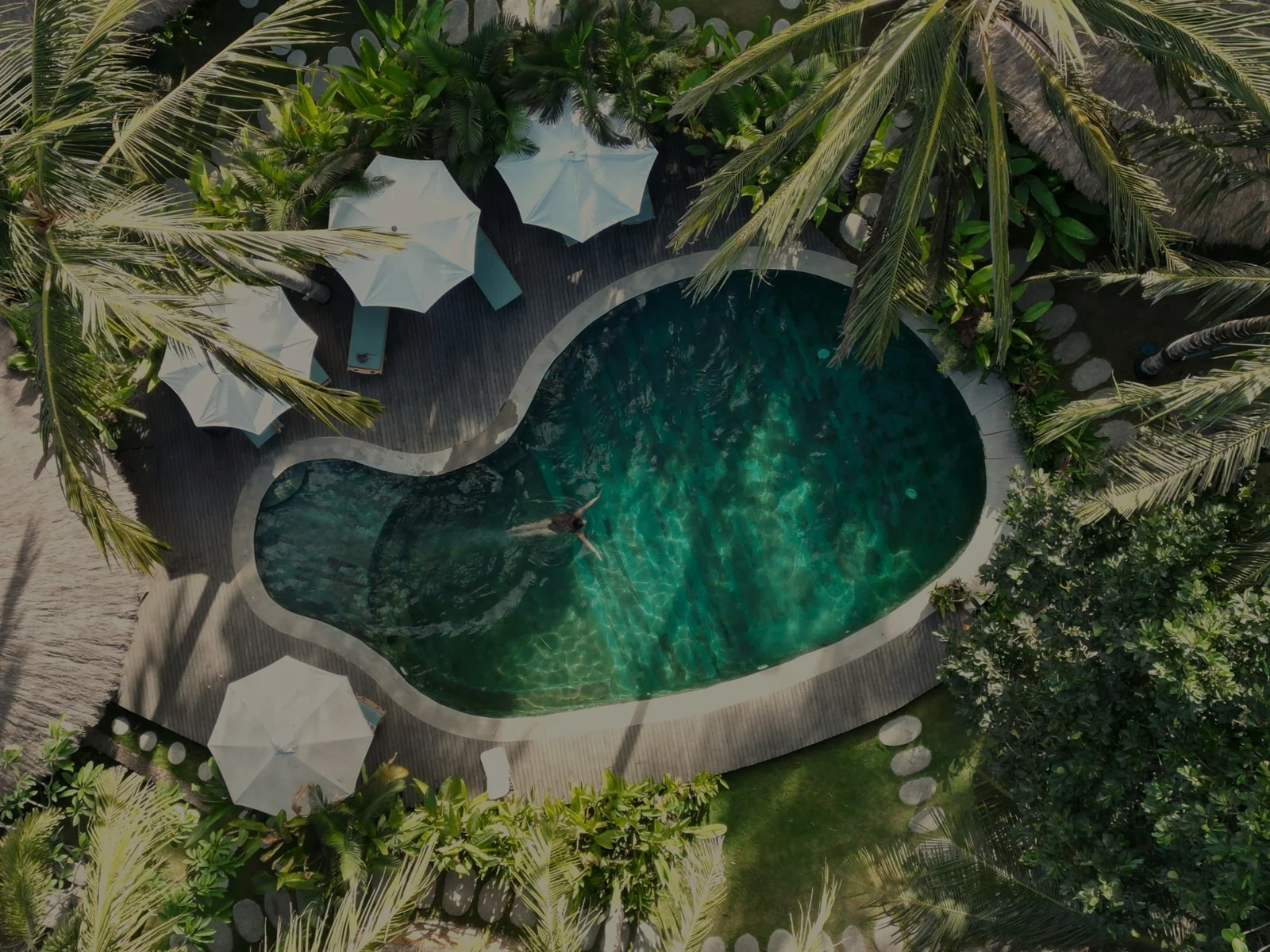 Aerial view of the lagoon pool at Mana Eco Retreat in Lombok, ringed by palms and white umbrellas