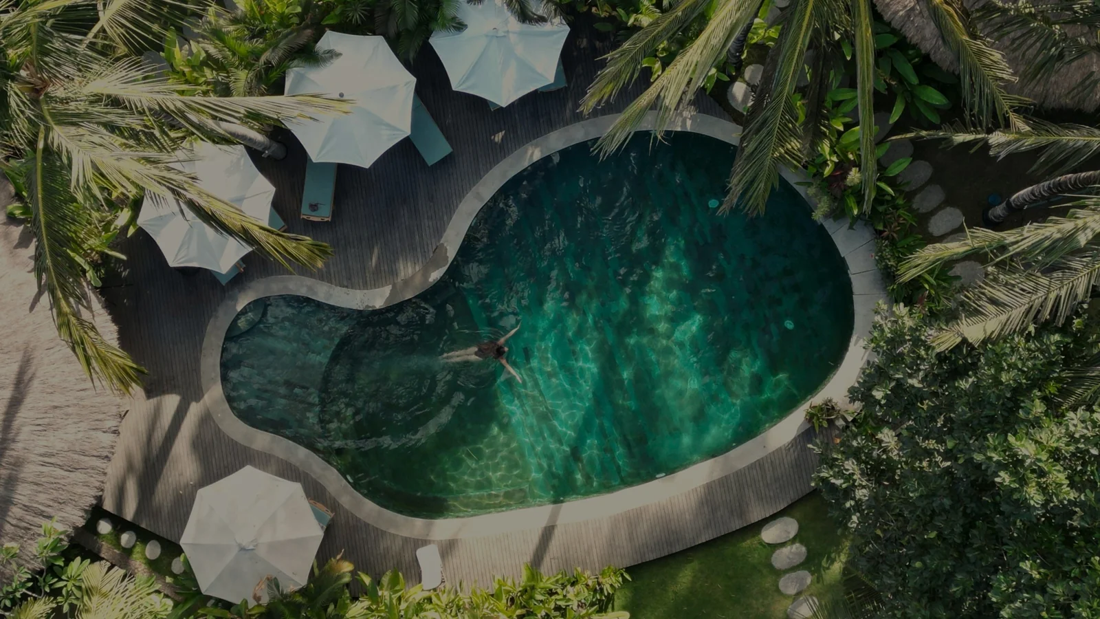 Aerial view of the lagoon pool at Mana Eco Retreat in Lombok, ringed by palms and white umbrellas